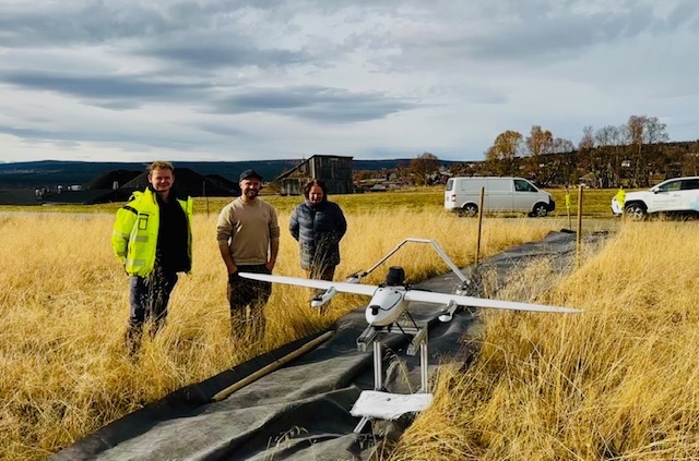 Three participants of the Røros-project standing in a field behind a drone